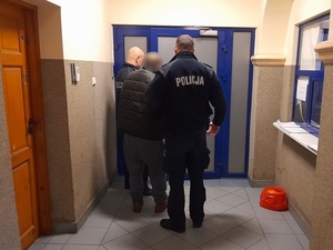 przed drzwiami pomieszczenia dla osób zatrzymanych stoją dwaj policjanci z zatrzymanym mężczyzną