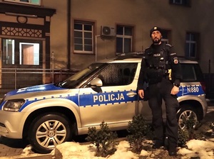 pora wieczorowa. Przed oznakowanym radiowozem stoi policjant służby patrolowej