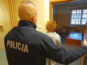 przed otwartymi drzwiami celi stoi kobieta. Za nią stoi policjant służby dyżurnej przytrzymując drzwi