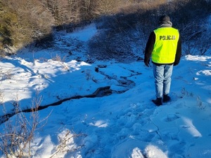 pora dzienna. Na ośnieżonym terenie przy wąskim strumieniu wody stoi policjant w kamizelce odblaskowej