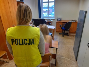 w pomieszczeniu służbowym policji na krześle siedzi zatrzymana kobieta. Za nią stoi policjantka w kamizelce odblaskowej z napisem policja