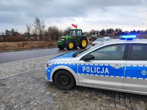 drogą jedzie ciągnik rolniczy z doczepioną flagą z barwami Polski. Na poboczu stoi policyjny radiowóz