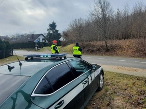 przy drodze stoją policjant i żandarm wojskowy. Za nimi znajduje się samochód Żandarmerii Wojskowej