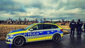 przy drodze obok oznakowanego radiowozu stoją policjanci. Drogą przejeżdżają ciągniki rolnicze