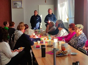 w sali przy długim stole siedzą seniorki i seniorzy. Przed nimi stoją policjant i policjantka