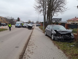 miejsce wypadku drogowego. Na jezdni stoi samochód osobowy z rozbitym przodem. Za nim znajduje się policyjny radiowóz. Z powodu wycieków płynów eksploatacyjnych jezdnia jest zasypana substancją neutralizującą. Kilka metrów przed tym samochodem, na pasie zieleni znajduje się drugi samochód osobowy z podobnymi uszkodzeniami. Jezdnią idzie policjant ruchu drogowego