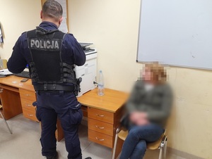 w policyjnym pomieszczeniu służbowym na krześle siedzi kobieta z rękoma skutymi kajdankami. Obok niej przy kserokopiarce stoi policjant