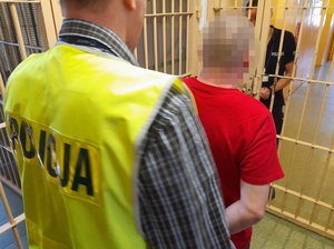 w okratowanym pomieszczeniu dla osób zatrzymanych policjant w kamizelce odblaskowej z napisem policja zakłada mężczyźnie kajdanki na ręce