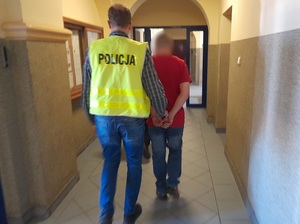 policjant w kamizelce odblaskowej z napisem policja prowadzi korytarzem mężczyznę skutego kajdankami