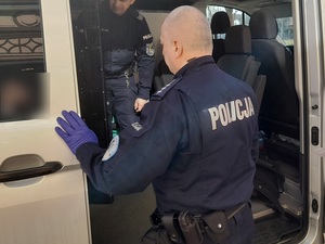 policjant służby konwojowej w rękawiczkach ochronnych przed otwartymi drzwiami nieoznakowanego radiowozu. Wewnątrz stoi drugi policjant