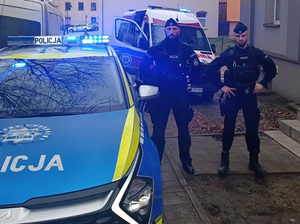 przy zaparkowanym przed budynkiem radiowozie z włączonymi sygnałami błyskowymi stoją dwaj policjanci służby patrolowej. Za nimi znajduje się karetka pogotowia