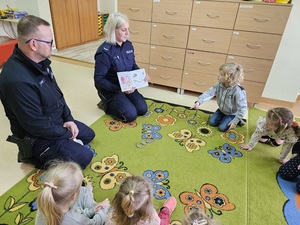 w sali przedszkola na dywanie siedzą dzieci. Przed nimi znajduje się dwoje policjantów. Policjantka trzyma otwartą książkę tekstem skierowaną w stronę dzieci