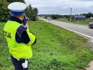 na poboczu drogi stoi policjant ruchu drogowego, który mierzy prędkość przejeżdżających pojazdów za pomocą ręcznego miernika prędkości