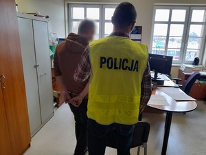 w pomieszczeniu służbowym nieumundurowany policjant w kamizelce odblaskowej z napisem policja trzyma za przedramię mężczyznę, który ma ręce skute z tyłu kajdankami