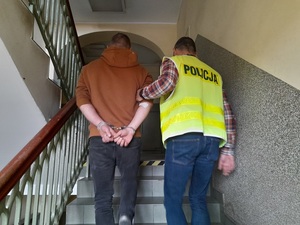 policjant w kamizelce odblaskowej z napisem policja prowadzi schodami w górę mężczyznę, który ma ręce skute z tyłu kajdankami