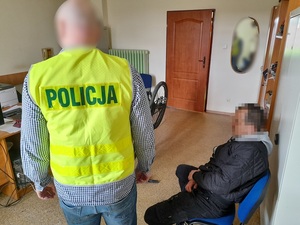 w policyjnym pomieszczeniu służbowym na krześle siedzi mężczyzna. Obok niego stoi policjant w kamizelce odblaskowej z napisem policja