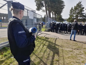przed ogrodzeniem stadionu stoją policjanci w kaskach i kamizelkach ochronnych. Wśród nich są kibice. Na pierwszym planie znajduje się policjantka w kamizelce ochronnej, trzymająca megafon