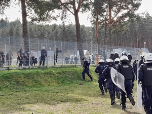 znajdujący się na stadionie pseudokibice próbują sforsować ogrodzenie. W ich stronę biegną policjanci z pałkami służbowymi, ubrani w kamizelki i kaski ochronne