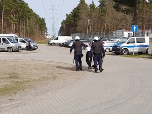 dwóch policjantów ubranych w kamizelki i kaski ochronne prowadzi w kierunku nieoznakowanego radiowozu zatrzymanego mężczyznę