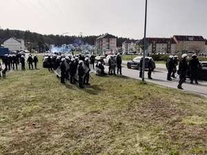 na pasie zieleni pomiędzy stadionem i jezdnią stoi kilkudziesięciu policjantów ubranych w kamizelki i kaski ochronne, z tarczami i pałkami służbowymi. Kilku policjantów stoi na jezdni przed samochodami. W tle widać chmurę dymu. Dwaj policjanci klęczą przy leżącym na ziemi mężczyźnie