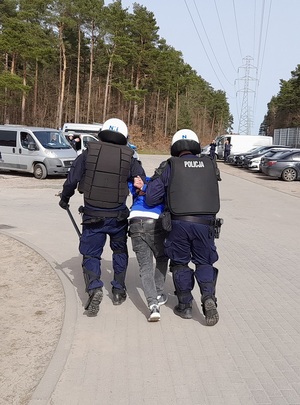 dwóch policjantów ubranych w kamizelki i kaski ochronne prowadzi w kierunku nieoznakowanego radiowozu zatrzymanego mężczyznę