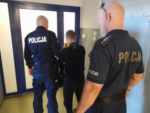Policjant prowadzi korytarzem komendy Policji zatrzymanego mężczyznę. Za nimi idzie drugi policjant