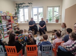 w bibliotece przed grupą przedszkolaków i opiekunek siedzi dwoje policjantów. Policjant czyta dzieciom książkę