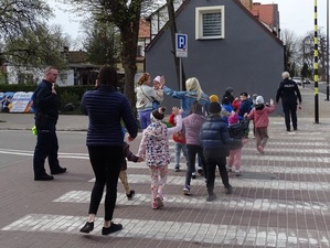 przez przejście dla pieszych przechodzi grupa przedszkolaków z opiekunkami. Każde dziecko ma rękę uniesioną w górze. Obok idzie policjant z ręką uniesioną w górze. Na czele idzie policjantka trzymająca tarczę do zatrzymywania pojazdów