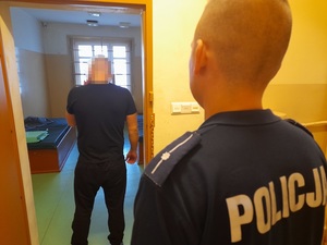 w otwartych drzwiach celi stoi skierowany tyłem do obiektywu mężczyzna. Za nim stoi policjant służby dyżurnej