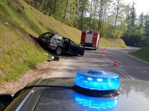 miejsce wypadku drogowego. Na łuku drogi na poboczu znajduje się rozbity samochód. Przed nim stoi wóz straży pożarnej, za nim policyjny radiowóz