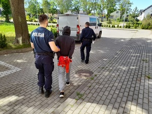 policjant prowadzi zatrzymanego mężczyznę w kierunku nieoznakowanego radiowozu. Przed nimi idzie drugi policjant