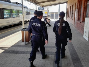 patrol składający się z trojga policjantów: dwóch policjantów i policjantki idzie peronem przy budynku dworca PKP. Na torach stoi pociąg, na ławce siedzi mężczyzna czytający gazetę