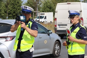 na ulicy stoją policjant i policjantka. Policjant mierzy prędkość pojazdów ręcznym miernikiem prędkości, policjantka trzyma tarczę do zatrzymywania pojazdów