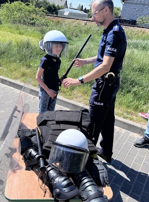 policjant pomaga chłopcu założyć policyjny sprzęt ochronny. Chłopiec ma na głowie policyjny kask, w ręce trzyma pałkę