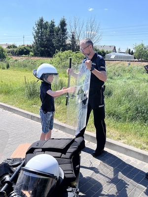 policjant podaje chłopcu tarczę oraz pałkę. Chłopiec ma na głowie policyjny kask wykorzystywany przy zabezpieczaniu imprez masowych