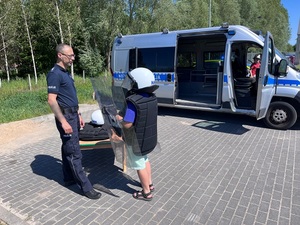 na parkingu przy oznakowanym policyjnym radiowozie policjant rozmawia z chłopcem ubranym w sprzęt ochronny wykorzystywany przy zabezpieczaniu imprez masowych