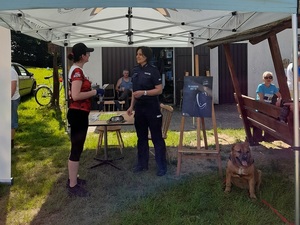 pod namiotem przy stoisku policyjnym stoją naprzeciw siebie  policjantka i  kobieta w stroju sportowym. Kobieta trzyma w dłoni ulotkę. Obok siedzi pies. Po prawej stronie stoiska na sztaludze jest zawieszony plakat ze zdjęciem policjanta i napisem zostań pomorskim policjantem