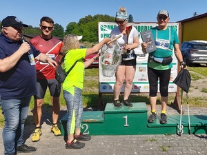 na podium z nr 1  stoi kobieta w stroju sportowym. Obok na podium na nr 3 stoi starszy mężczyzna w stroju sportowym i kijkami do nordic walking, trzymający książkę. Kobieta na podium otrzymuje od innej kobiety kosz z prezentami. Obok stoi mężczyzna w stroju sportowym oraz starszy mężczyzna z mikrofonem w dłoni