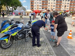 na placu policjantka zakłada kilkulatkowi na rękę opaskę odblaskową. Obok stoi policyjny oznakowany motocykl