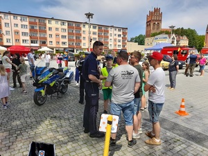 na placu policjant rozmawia z grupą osób. Obok stoi policyjny oznakowany motocykl