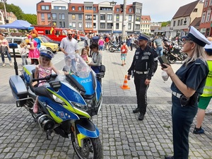 na placu na policyjnym oznakowanym motocyklu siedzi kilkulatka. Inspektorka ITD robi jej zdjęcie. Obok stoi policjant ruchu drogowego w kurtce motocyklowej. W tle widać przemieszczających się placem ludzi