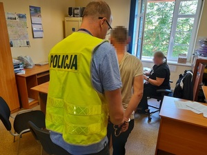 w pokoju przesłuchań policjant w kamizelce odblaskowej z napisem policja zdejmuje mężczyźnie z rąk kajdanki. Przy biurku siedzi nieumundurowana policjantka