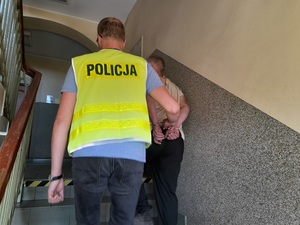 policjant w kamizelce odblaskowej z napisem policja prowadzi schodami w górę mężczyznę z rękoma skutymi z tyłu kajdankami