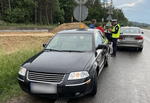 na poboczu stoi czarny vw passat. Przy nim stoi policjant ruchu drogowego oraz kobieta i mężczyzna. Dalej zaparkowany jest nieoznakowany policyjny radiowóz