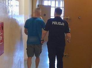 korytarzem pomieszczenia dla osób zatrzymanych, w kierunku otwartych drzwi celi idą policjant i zatrzymany mężczyzna