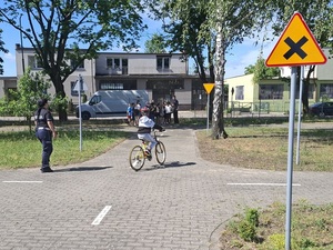 miasteczko ruchu drogowego. Policjantka ruchu drogowego obserwuje chłopca jadącego rowerem. Na pierwszym planie znajduje się znak drogowy informujący o skrzyżowaniu równorzędnym