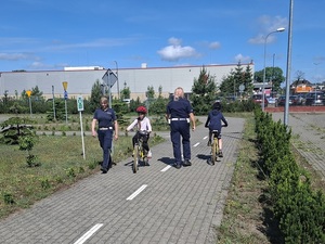 miasteczko ruchu drogowego. Na drodze wymijają się dwie dziewczynki jadące rowerami. Obok każdej z nich idą policjantki ruchu drogowego