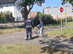 miasteczko ruchu drogowego. Przed skrzyżowaniem oznaczonym znakiem stop znajduje się chłopiec na rowerze. Obok stoi policjant ruchu drogowego