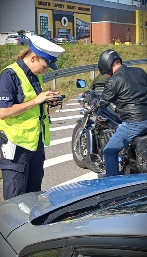 na poboczu policjantka ruchu drogowego wpisuje dane do urządzenia elektronicznego. Obok na motocyklu siedzi mężczyzna