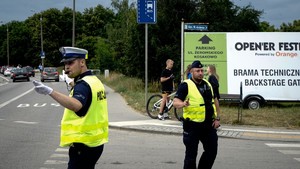 Policjant ruchu drogowego z gwizdkiem w ustach kieruje ruchem na skrzyżowaniu. Za nim obok przejścia dla pieszych stoi policjant służby prewencyjnej. Za nim znajduje się baner festiwalu open`er